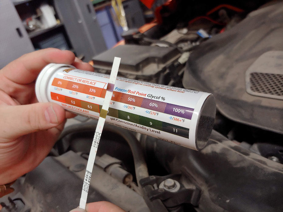 The image depicts a person holding a package of coolant and brake fluid test strips in front of a car engine. Text on the package displays a chart measuring freeze and boiling points, a glycol percentage scale, and a pH corrosion level.