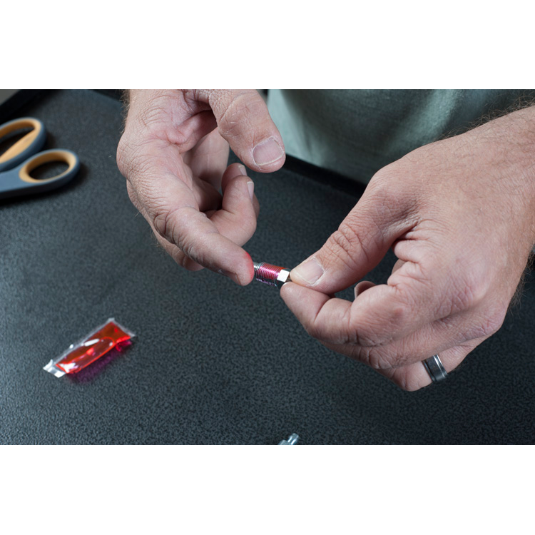 The image is a close-up of a man’s hands holding a small, round object with a hole in the center. The object is most likely a brake fluid bleeder screw cap and putting red grease on it