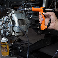 The image shows a mechanic using a brake fluid bleeding gun to bleed a brake caliper on a car.